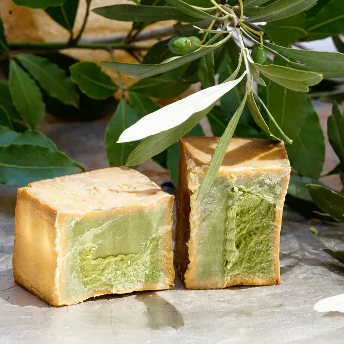 Two rectangular bars of handmade soap with a light brown outer layer and a green marbled center, displayed on a stone surface with olive branches in the background.