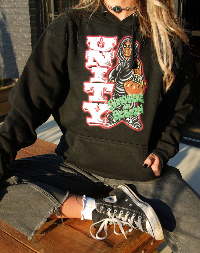 A person sits cross-legged wearing a custom UNITYTX black hoodie by Independent Trading Co., featuring a vibrant graphic of a woman with a basketball and bold UNITY text, paired with gray jeans and black Converse sneakers.