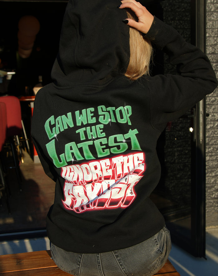 A person with blonde hair, wearing a black UNITYTX printed sweatshirt with colorful text on the back reading CAN WE STOP THE LATEST UNDER THE RADAR, sits outdoors on a wooden bench facing away from the camera.