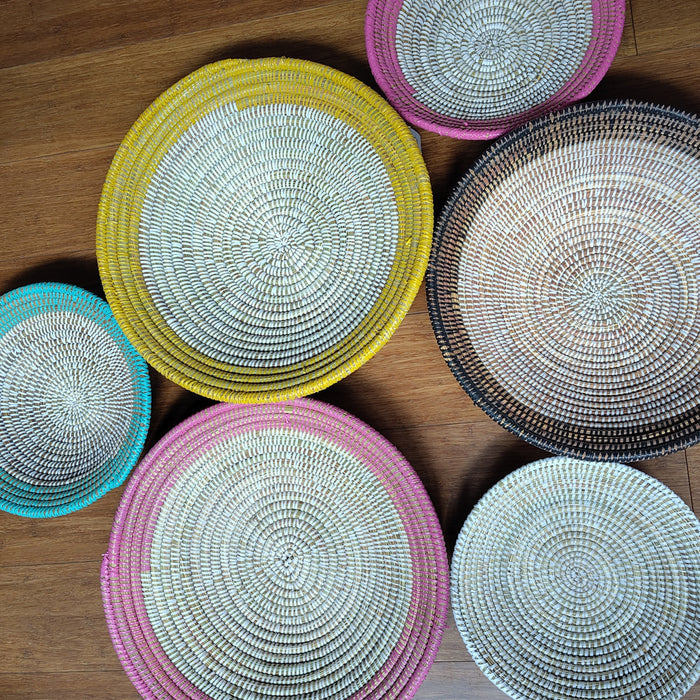 Six round woven basket bowls in various colors and sizes arranged on a wooden floor. The bowls feature a spiral woven pattern with rims in pink, yellow, teal, black, and white, set against a warm brown wood background.