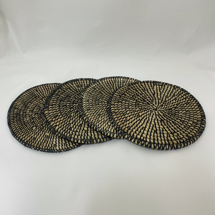 Four round woven seagrass placemats with a black and natural beige spiral pattern, arranged in a fan shape on a white fabric background.
