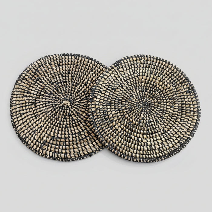 A set of two round placemats with a geometric pattern, featuring a mix of black and beige river grass with white binding.