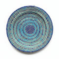 A round woven basket plate with concentric spiral patterns in blue, turquoise, and brown, displayed on a plain white background.