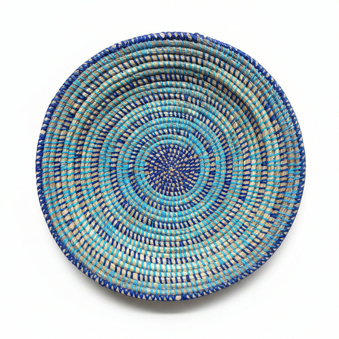 A round woven basket plate with concentric spiral patterns in blue, turquoise, and brown, displayed on a plain white background.