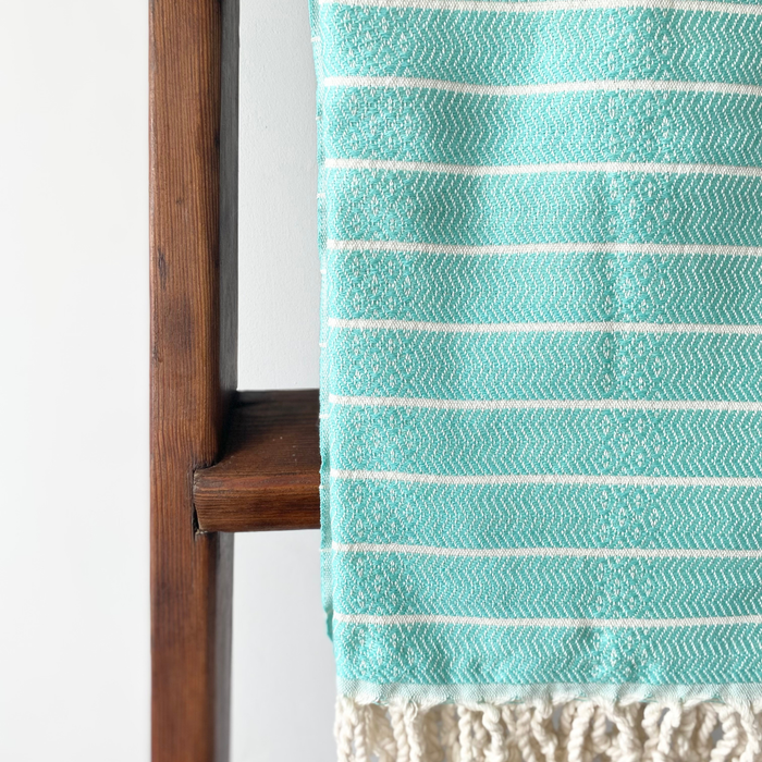 A close-up of a Turkish towel in mint green and white, draped over a dark wooden stand against a plain light background. The fabric shows a striped pattern with subtle geometric and floral motifs, and a cream-colored fringed edge is visible at the bottom.