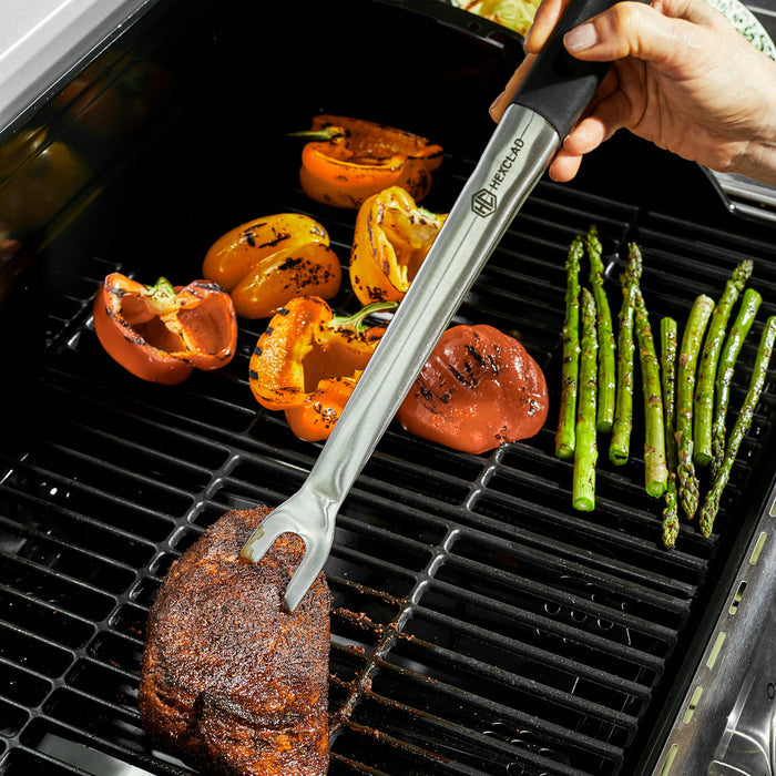 HexClad meat prongs poking at a piece of meat on a grill. There are also vegetables on the grill.