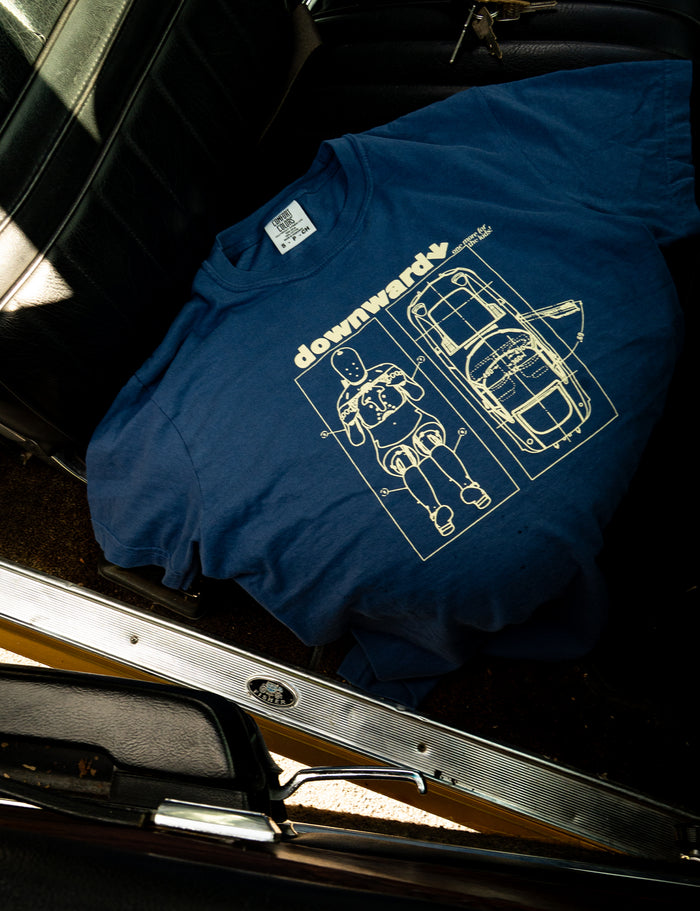 A blue DOWNWARD Comfort Colors printed shirt, featuring a downward graphic and a crash test dummy illustration, is laid out on the black leather seat of a car.