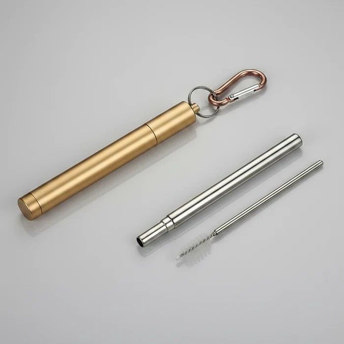 A gold colour metal folding straw caseshown on a grey surface, including   a carabiner clip and  a long metal straw and a small brush.