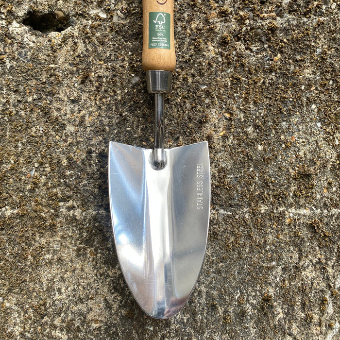 A small hand trowel with a polished steel blade and a light wood handle, resting on a textured concrete surface.