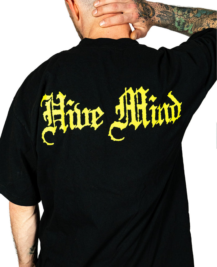 A person with tattooed arms and a shaved head faces away, wearing a spacious fit black Knocked Loose T-shirt featuring Hive Mind in large yellow Gothic letters on the back. Made from shrink-free cotton for lasting comfort.