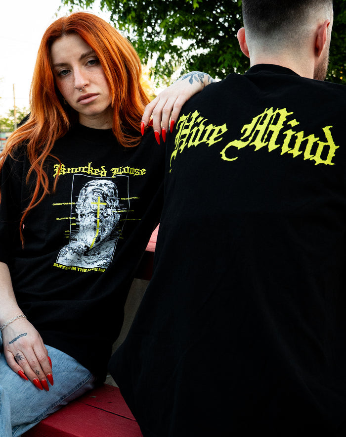 Two people sit outdoors on a red bench. The woman with long red hair and tattoos wears a black shirt with yellow graphics. The man, back turned, wears a Knocked Loose LA Apparel T-Shirt with “Hive Mind” in yellow gothic text. Green trees are behind them.