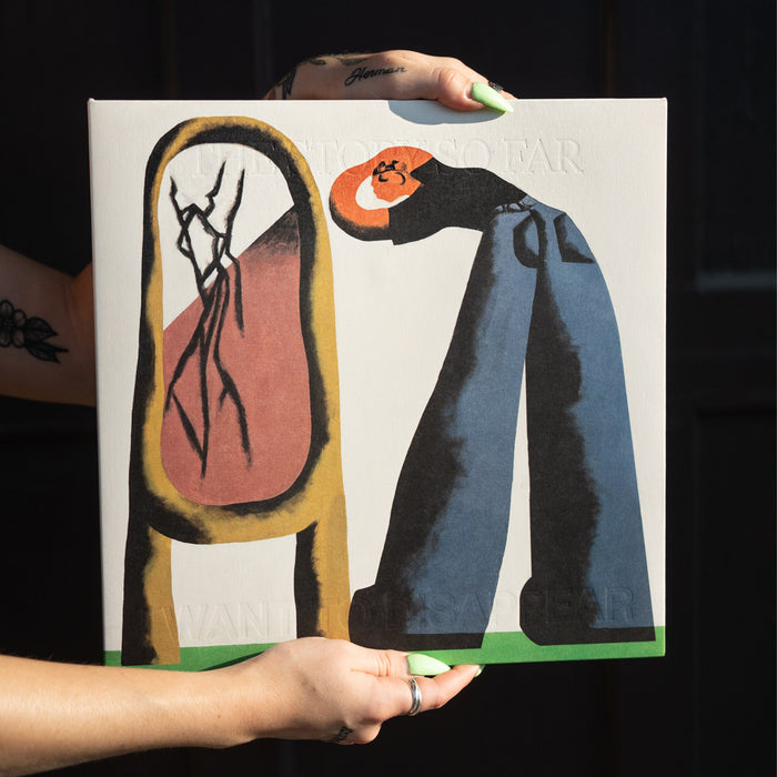 A person holds a limited edition 4th Press Milky Clear Rainbow Splatter vinyl album by The Story So Far, featuring abstract artwork with a cracked mirror and a figure wearing blue pants, black shoes, and an orange face set against a green and white background.