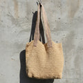 A natural beige woven tote bag with dual shoulder straps, hanging against a textured concrete wall in outdoor sunlight.