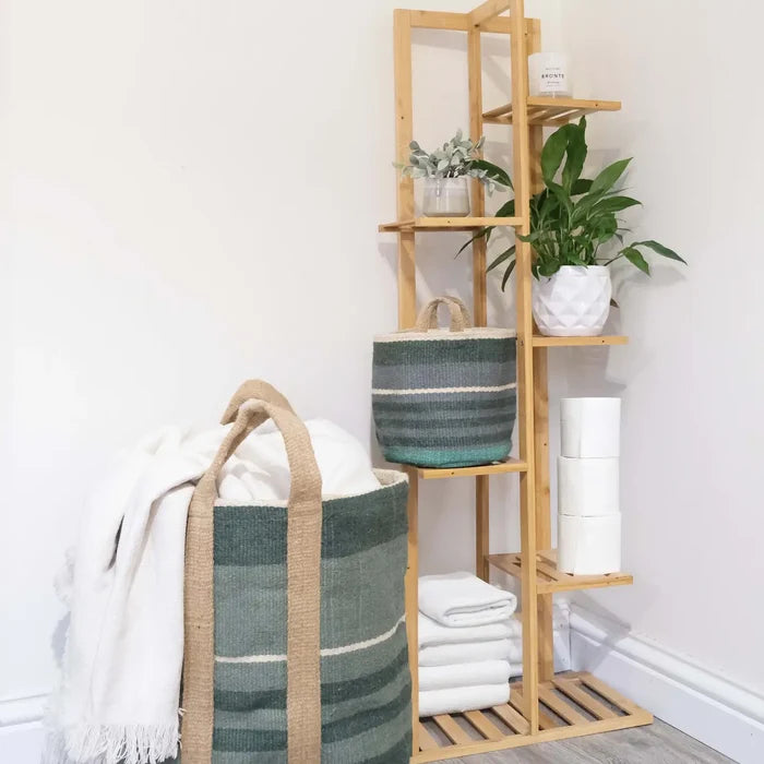 A tall light wood shelving unit styled with plants, a striped green and beige storage basket, rolled white towels, and a white throw blanket draped over a large matching woven basket on the floor. The scene is set against a plain white wall and light wood flooring.