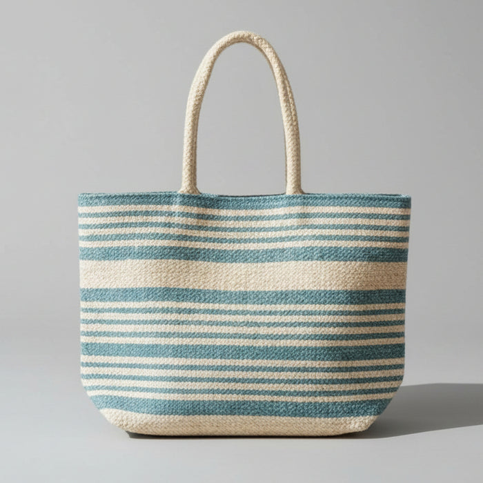 A striped woven tote bag with natural jute handles sits on a light wood floor in a bright, modern room with large windows and strong sunlight.
