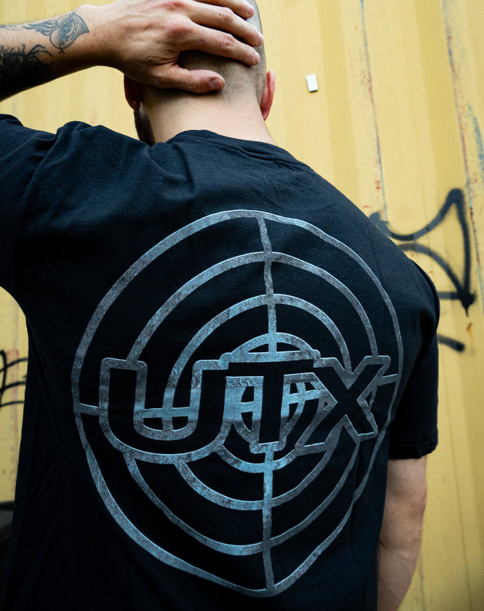 A tattooed individual stands before a yellow graffiti wall, hand on their shaved head, wearing a black UNITYTX Comfort Colors tee featuring a large circular target and "UTX" graphic printed on the back.