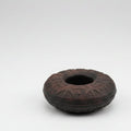 A dark-colored metal finger bell with embossed patterns and a central hole for wearing on a finger.