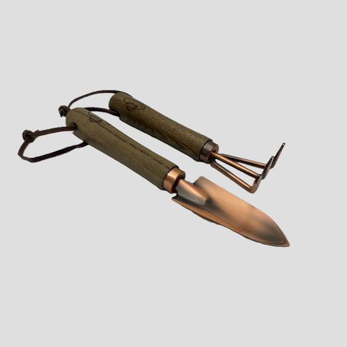 A pair of small hand tools with olive green wooden handles and copper-colored metal heads, including a trowel and a weeding fork, shown on a solid light gray background.