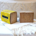 A bar of handmade Karwan soap displayed next to its yellow paper packaging box, resting on a white fringed textile against a white carved ceramic background.