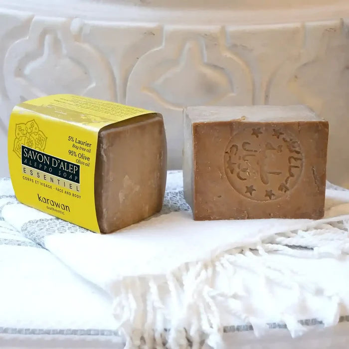 A bar of handmade Karwan soap displayed next to its yellow paper packaging box, resting on a white fringed textile against a white carved ceramic background.