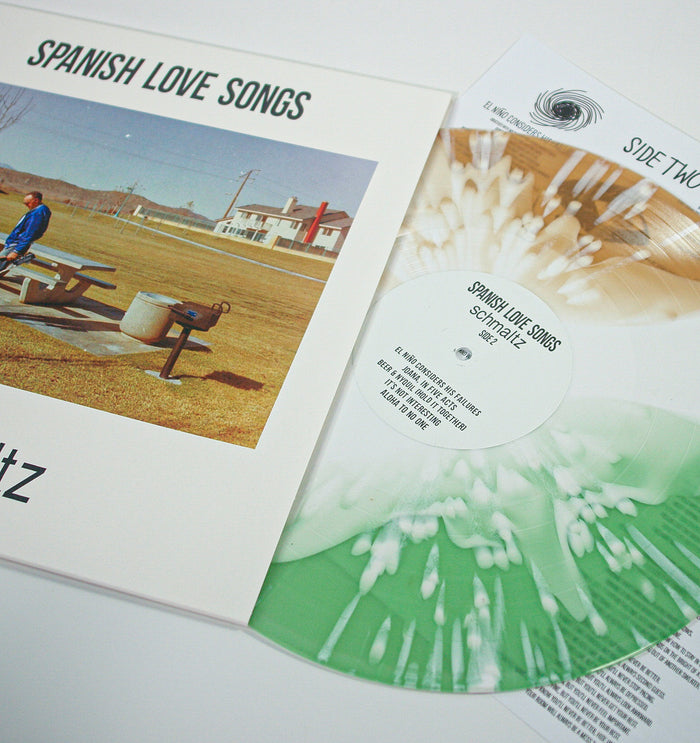 A green and white splatter vinyl record from Spanish Love Songs sits beside its cover, which shows a person at a picnic table with beer bottles in a grassy field. The album is titled "Schmaltz.