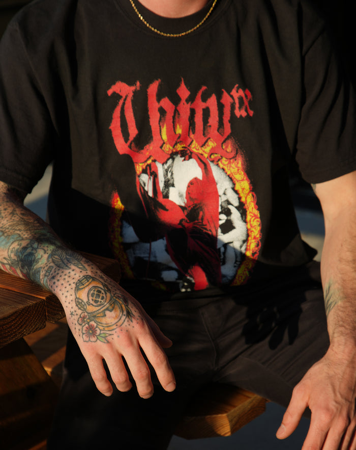 A person with tattooed hands sits on a wooden bench, wearing a black UNITYTX Comfort Colors t-shirt featuring a bold red and yellow graphic and the word "Thy" in stylized letters; their face is not visible.