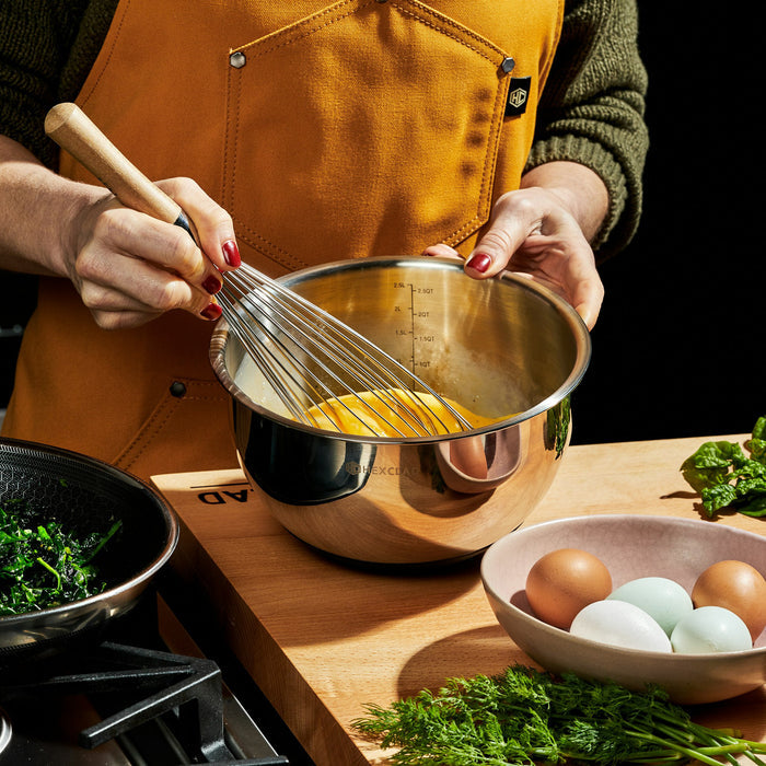 Someone with red painted finger nails whisking eggs in a HexClad stainless steel mixing bowl, with more eggs, spinach, and herbs on the counter.