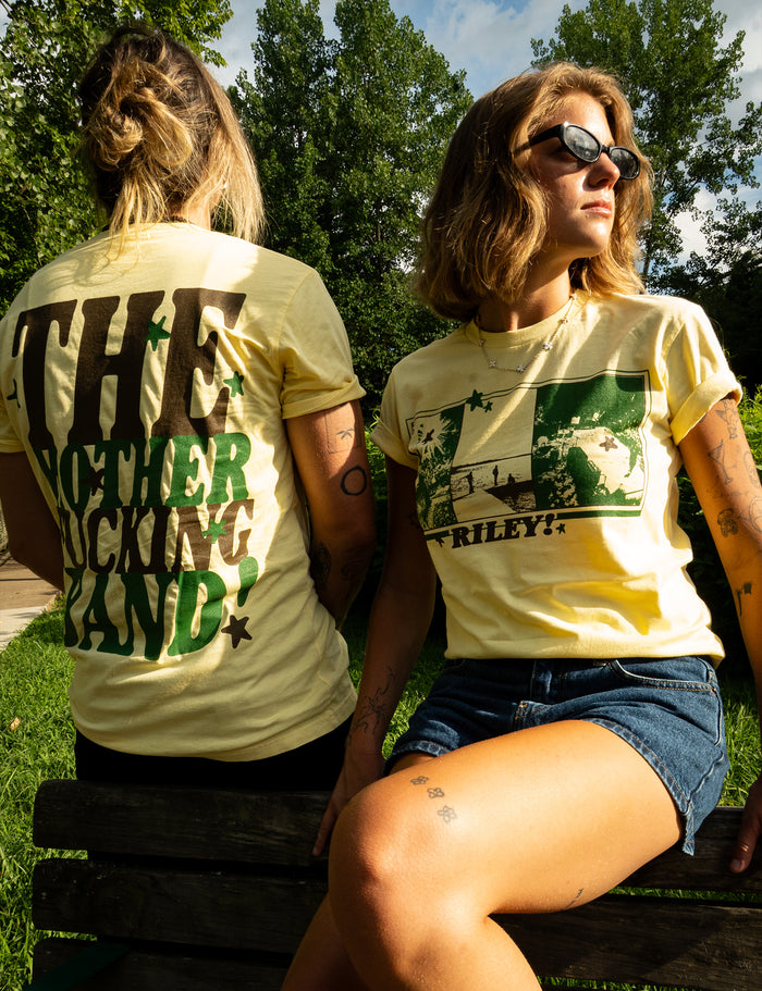 Two women wearing yellow Riley! Premium t-shirts sit on a park bench—one with her back turned, showing off bold green printed text, the other facing forward in sunglasses and shorts. Sunlight filters through the trees in the background.