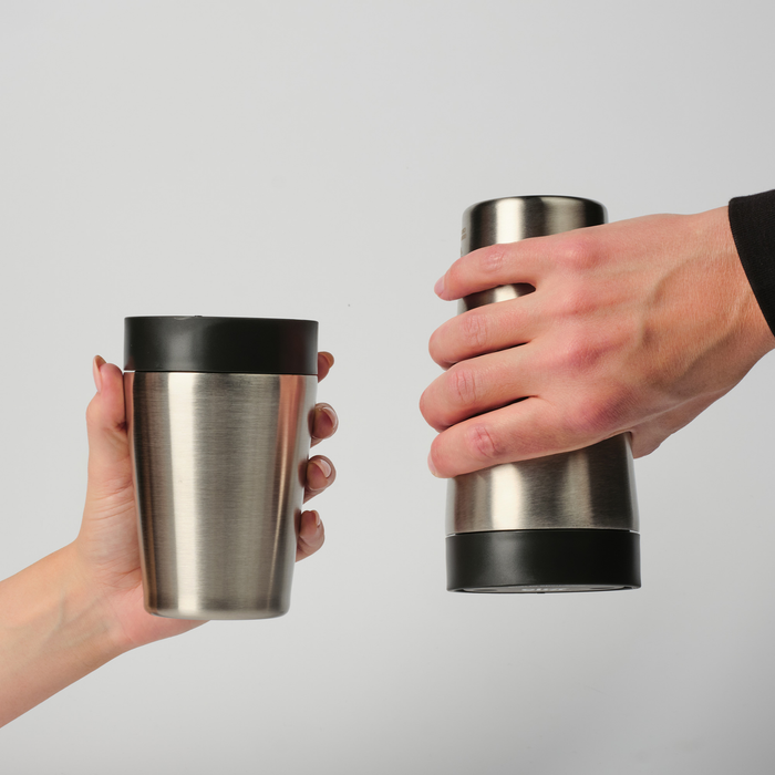 Two hands holding stainless steel travel tumblers with black lids against a light gray background.