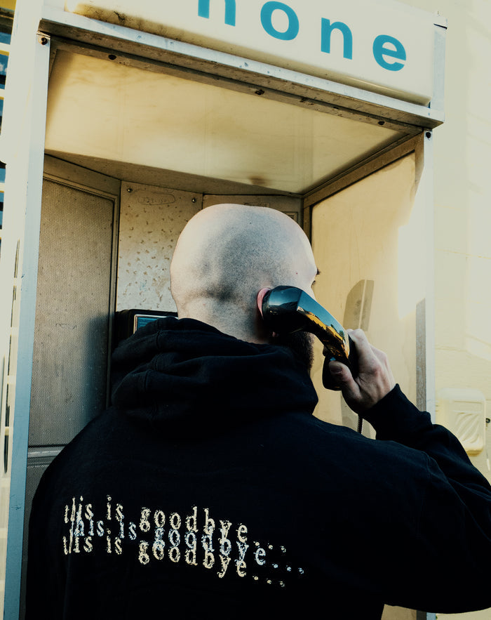 A bald person wearing a black printed Chamber sweatshirt stands in a phone booth, holding the receiver. "this is goodbye... this is goodbye..." is written on the back, while the word phone appears partly above.