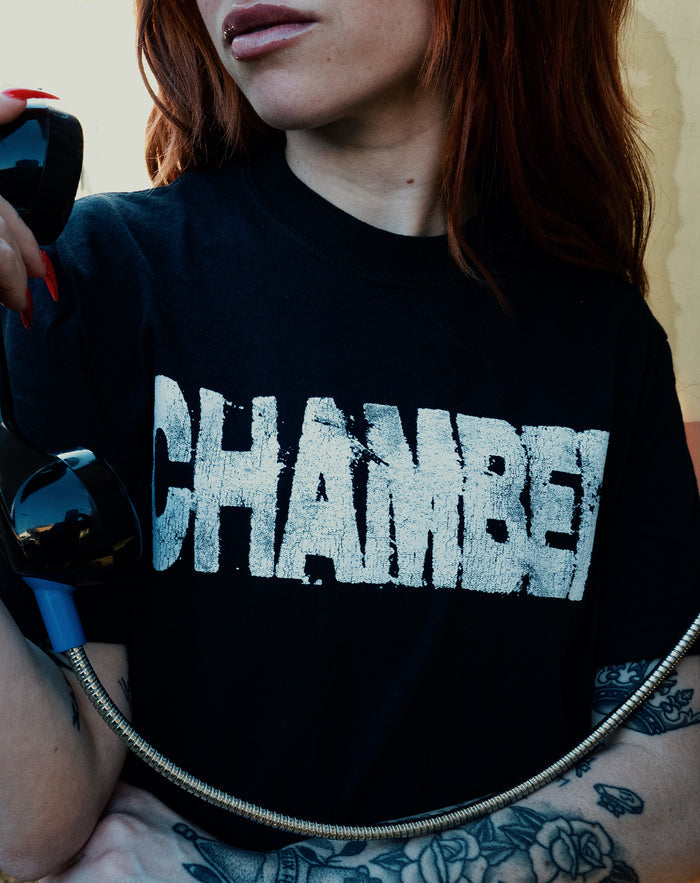 A red-haired, tattooed person wears a black Chamber t-shirt with bold, distressed white CHAMBER lettering, holding a black phone receiver with a metal cord.