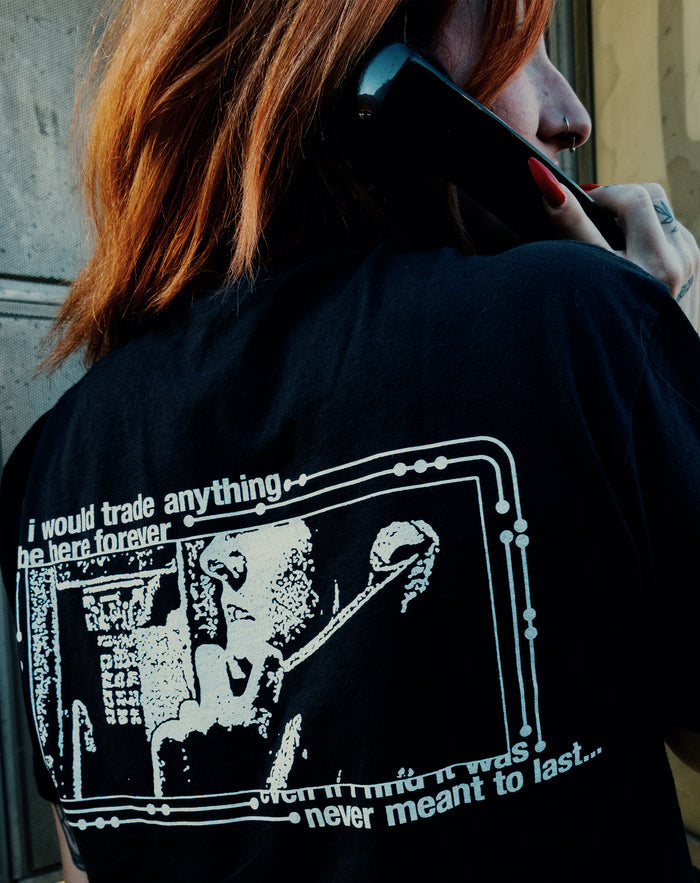 A red-haired person holds a phone to their ear, wearing a Chamber Comfort Colors t-shirt featuring white graphic art and partially visible text: "i would trade anything, ... be here forever ... never meant to last...".