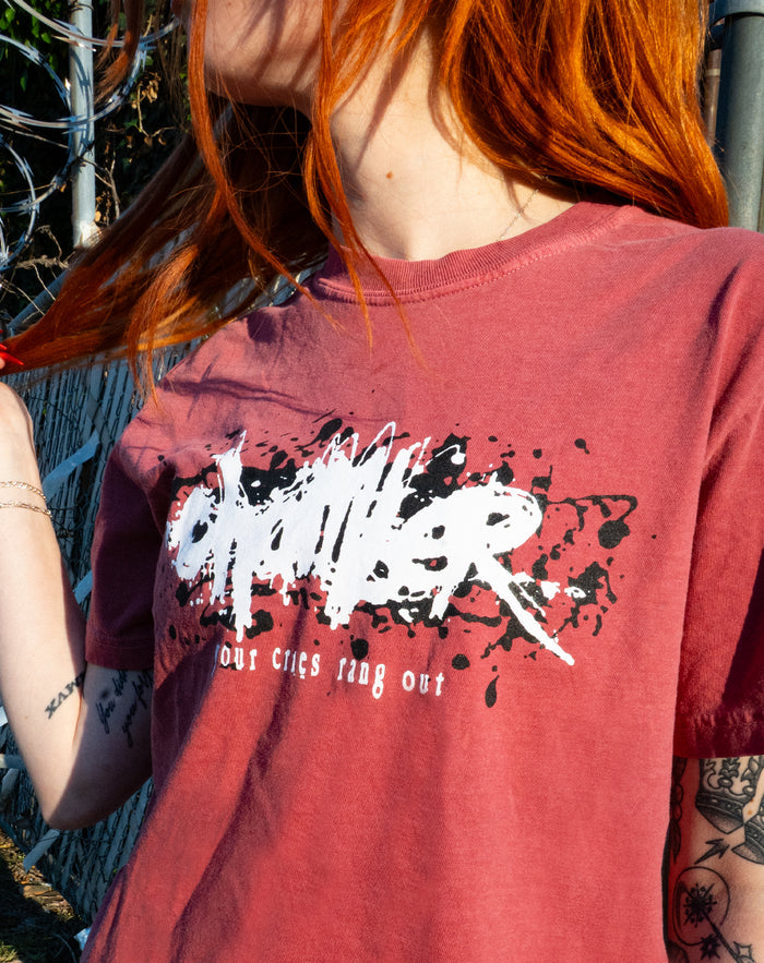 A person with long orange hair wears a faded red Chamber t-shirt printed with "CAPTIVER Our Cries Ring Out," standing by a barbed wire fence in bright sunlight.