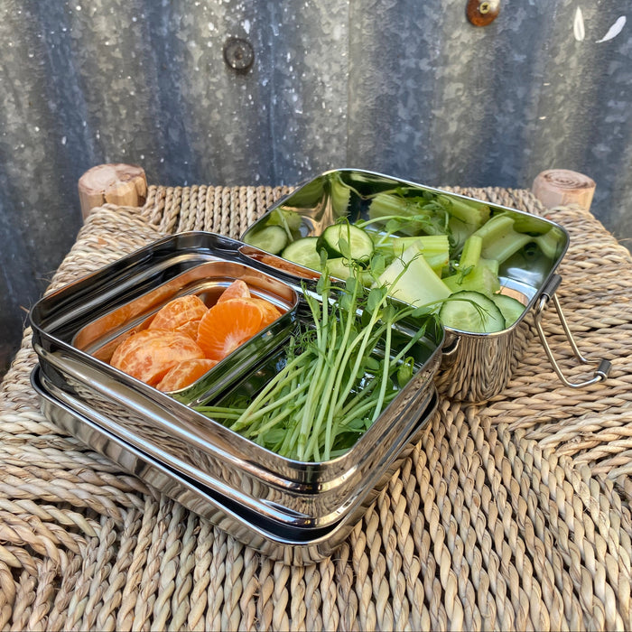 A stainless steel bento lunch box with three compartments, shown open and filled with sliced oranges, green herbs, sliced cucumbers, and avocado, placed on a woven wicker table against a corrugated metal wall.