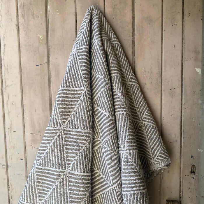 A woven throw blanket with a geometric striped pattern in taupe and off-white, hanging on a rustic wooden door.